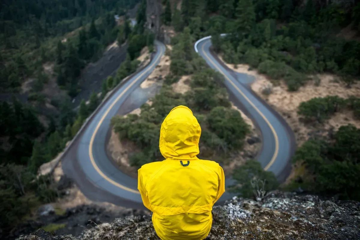 A person in a yellow jacket sitting on a cliff edge, overlooking a winding road splitting into different directions below